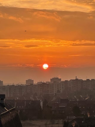 View of a city landscape at dusk with buildings in the foreground. Sky is yellow and red, with the sun visible in the center of image.