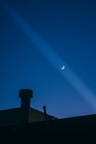 Dark blue night sky with a roof of a building and chimney on the foreground. A partial moon is visible in a beam of light.