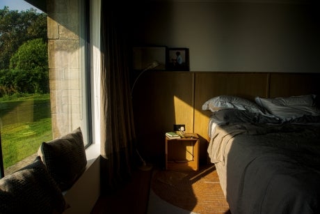 View into a bedroom, partially in shade. Bed on the right, window through which a green yard can be seen on the left.