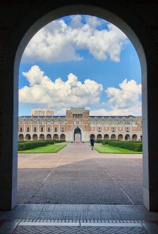 Lovett Hall shot through the archway in front of Fondren Library. Statue of Rice is visible between the buildings, with a lone figure walking towards it.