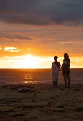 A couple with their backs to the camera stand in front of a sunset view over water.