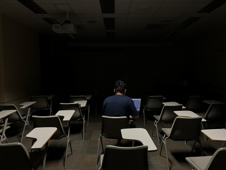 Person is sitting in a dark empty classroom with back to camera, working on a laptop.