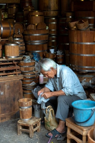 An elderly man sitting on a stool in his wood workshop on the streets of Thailand, hard at work not looking toward the camera.