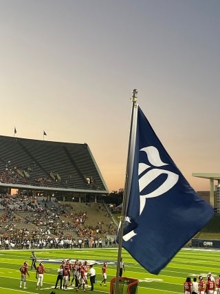 Football field with players standing in a group and spectators in the stands. Large Rice University flag on the foreground.