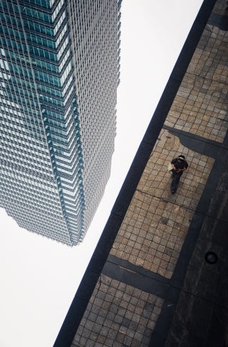 A skyscraper viewed upside down, descending from top left corner of image. Right side of picture shows a reflection of a sidewalk with a pedestrian.